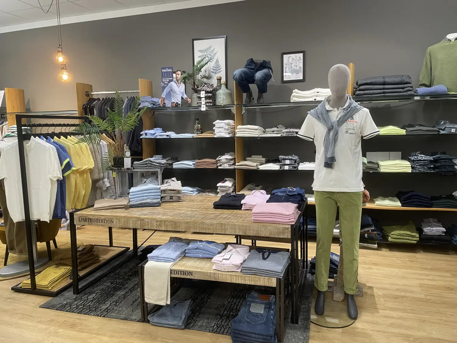 Abig-Moden - Modeboutique - Innenansicht eines hell erleuchteten Herrenbekleidungsgeschäfts mit einer Schaufensterpuppe in weißem Polohemd und grüner Hose, im Hintergrund ordentlich aufgereihte Regale mit gefalteten Hemden und Jeans.
