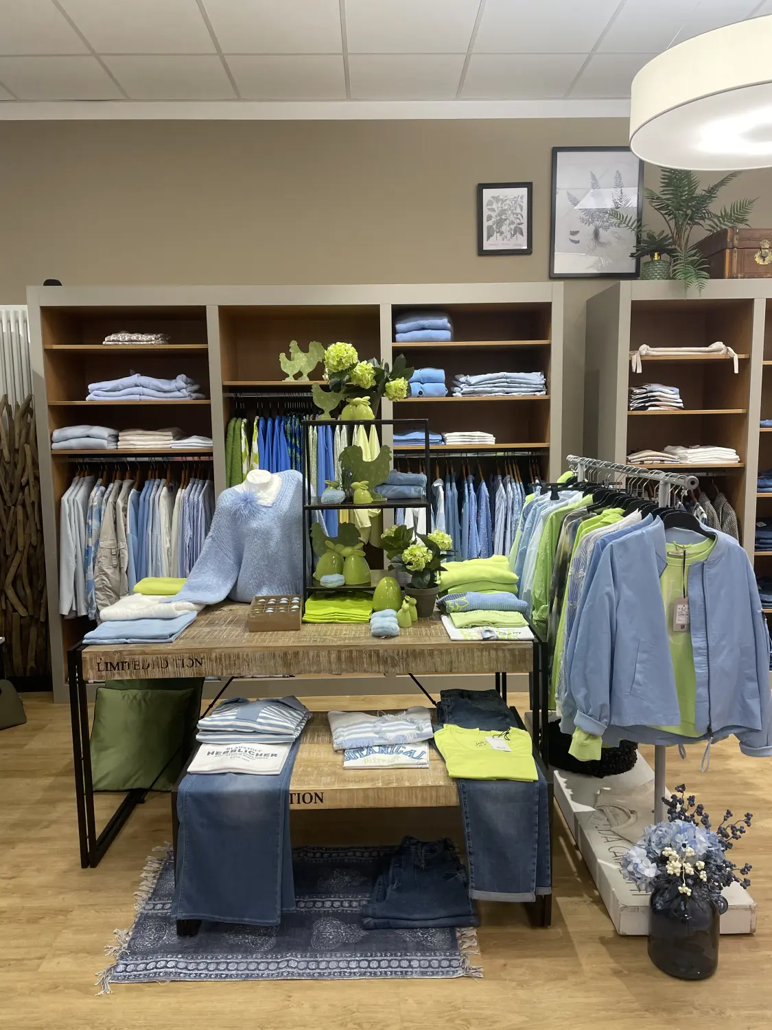 Abig-Moden - Modeboutique - In einer Boutique ist ein Ausstellungsbereich zu sehen, auf dem ein rustikaler Holztisch mit gefalteter blauer und gelber Kleidung liegt, flankiert von Regalen und Kleiderständern mit aufeinander abgestimmter Frühlingskleidung.
