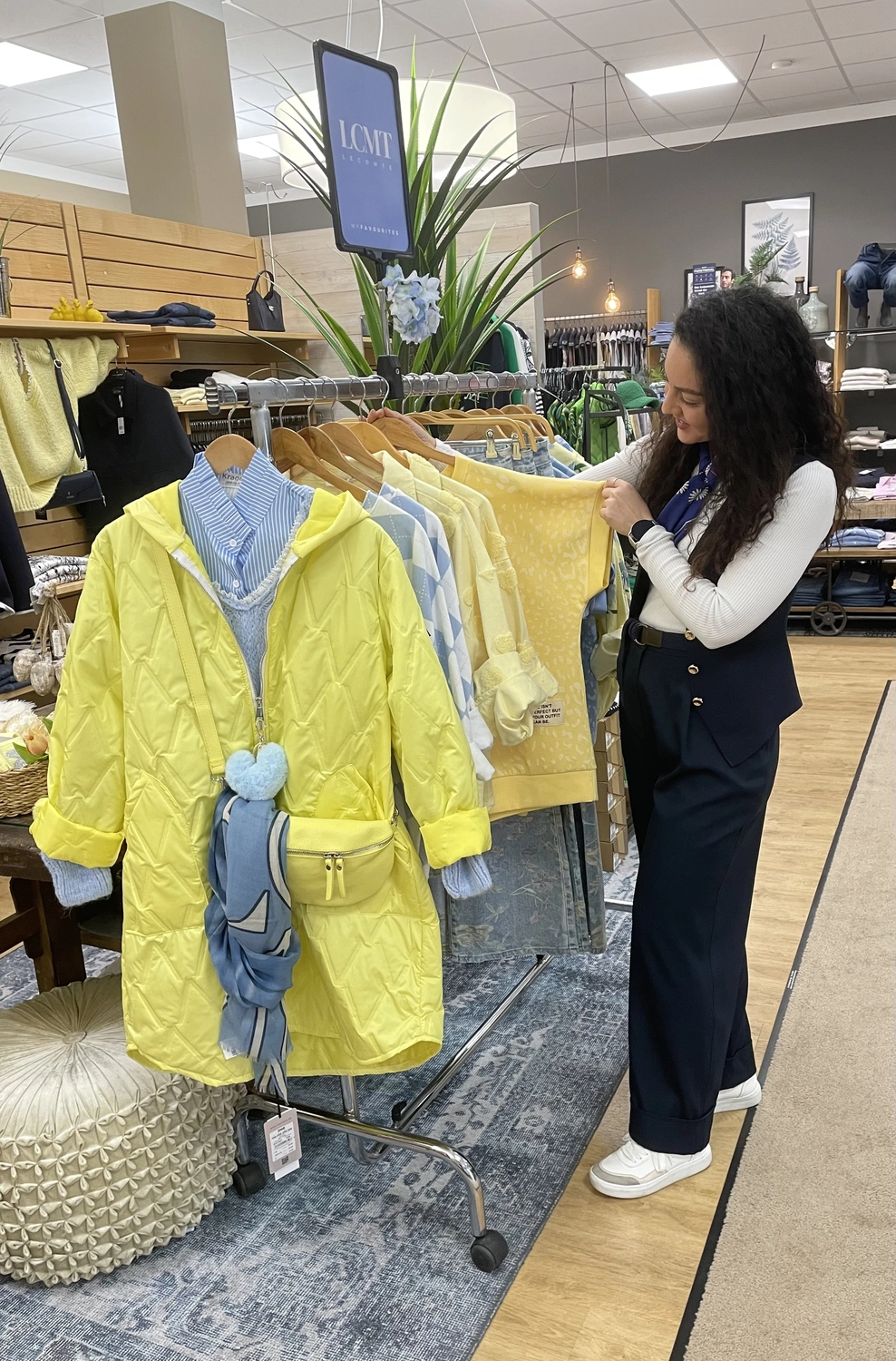 Abig-Moden - Modeboutique - Eine Frau stöbert in einem Geschäft an einem Kleiderständer und betrachtet dabei einen leuchtend gelben Steppmantel und ein gestreiftes blaues Hemd.