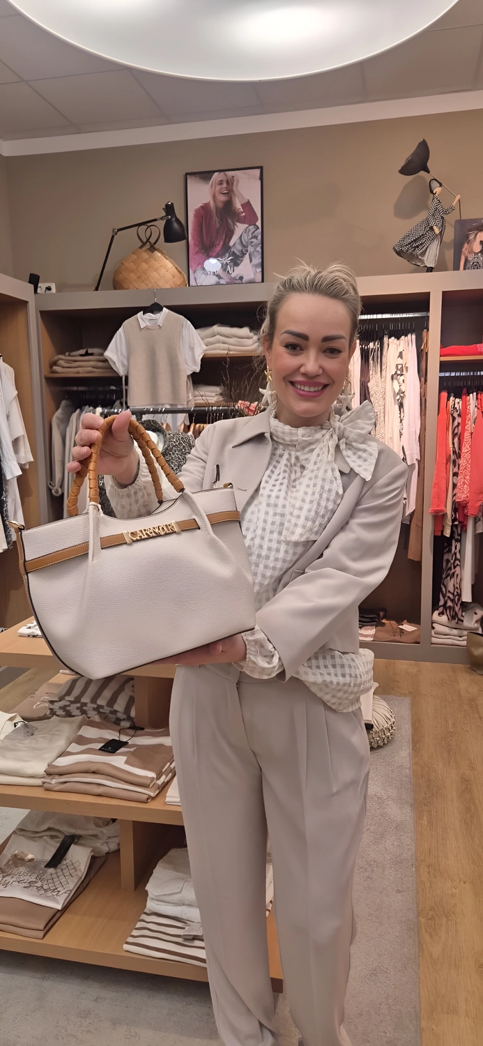 Abig-Moden - Modeboutique - Eine lächelnde Frau in einem beigen Blazer und einer karierten Bluse mit Schleife hält eine große weiße Handtasche mit hellbraunen Ledergriffen hoch.