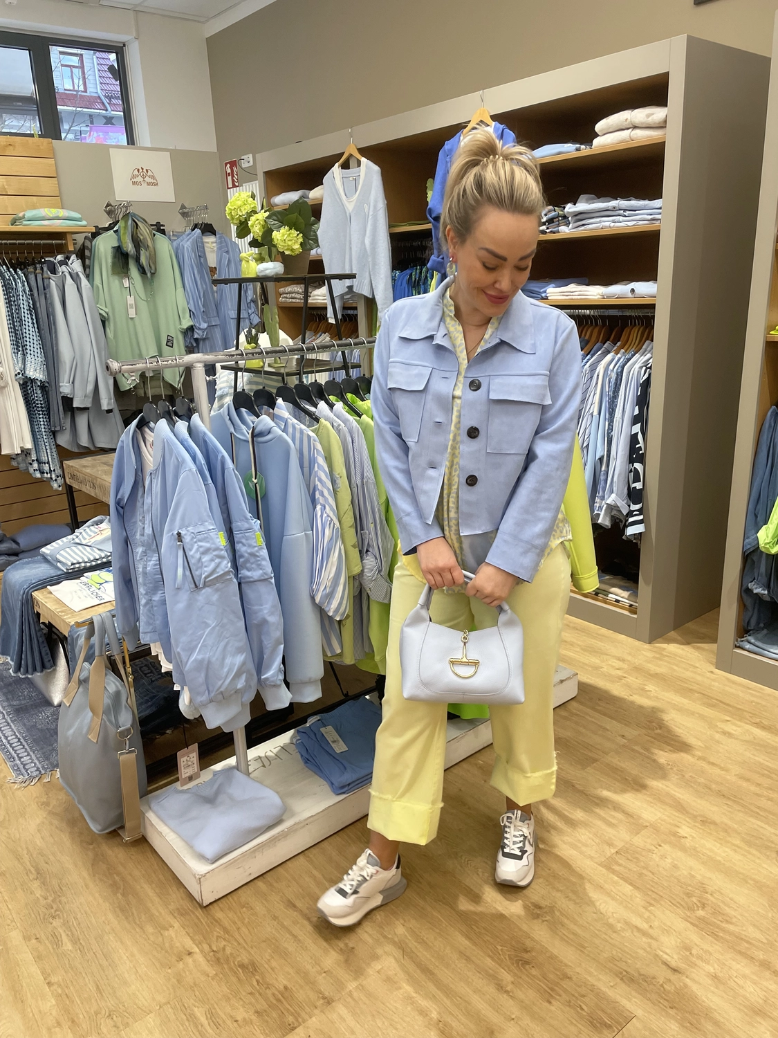Abig-Moden - Modeboutique - Eine Frau posiert in einer Bekleidungsboutique in einem hellblauen Hemd und einer gelben Hose, während sie eine weiße Handtasche hält.