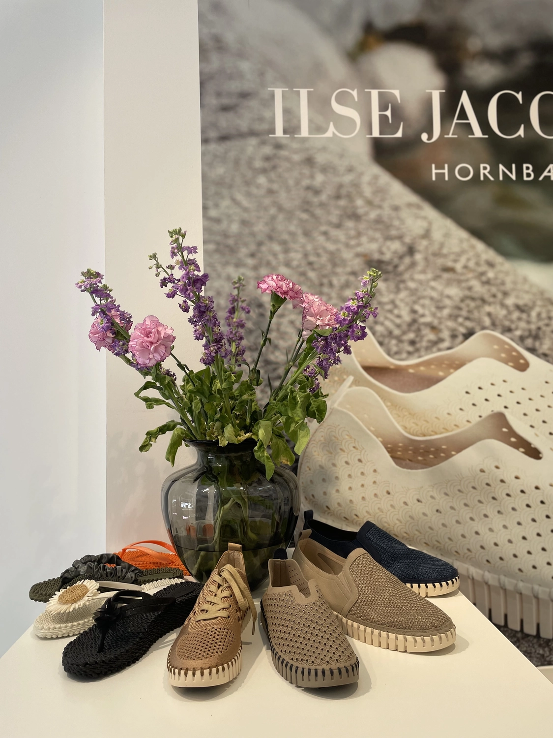 Abig-Moden - Modeboutique - Ein Verkaufsdisplay mit einem Strauß lila Blumen in einer Glasvase neben mehreren Paar gewebten Slippern und einem großen Ilse Jacobsen Hornbæk Markenplakat.