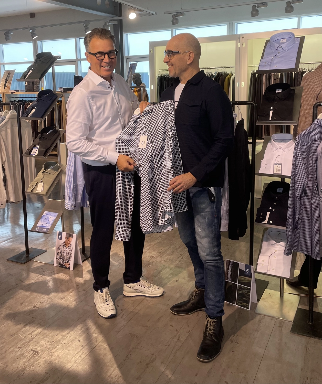 Abig-Moden - Modeboutique - Zwei Männer unterhalten sich in einer Boutique über ein gemustertes Hemd, das der Mann links hochhält.