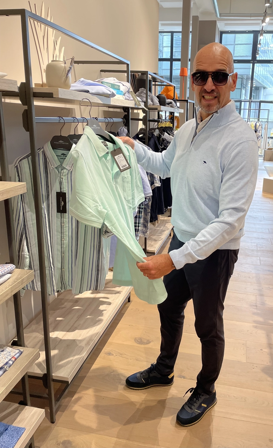 Abig-Moden - Modeboutique - Ein Mann mit Sonnenbrille lächelt und hält in einem Bekleidungsgeschäft ein hellgrünes Poloshirt in der Hand.