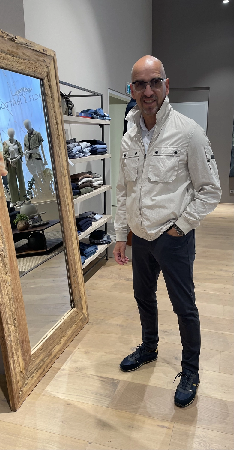 Abig-Moden - Modeboutique - Ein kahlköpfiger Mann mit Brille, beiger Funktionsjacke mit Reißverschluss und dunkler Hose posiert in einem hell erleuchteten Bekleidungsgeschäft.