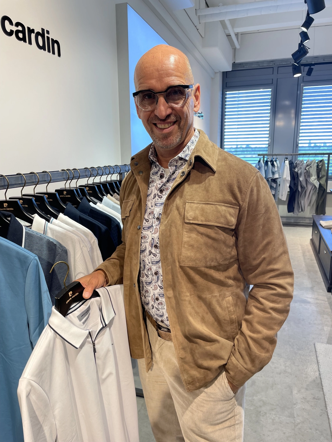 Abig-Moden - Modeboutique - Ein Mann mit Sonnenbrille lächelt und hält in einem Bekleidungsgeschäft ein hellgrünes Poloshirt in der Hand.