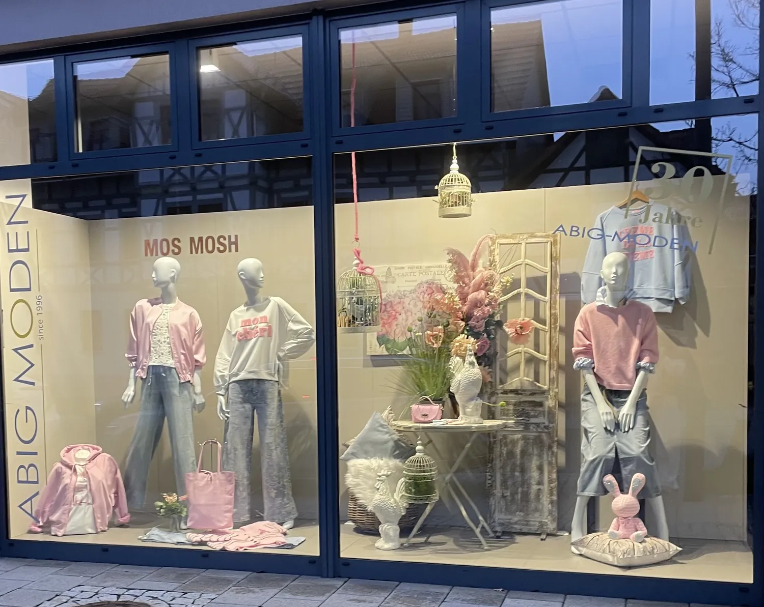 Abig-Moden - Modeboutique - Ein Schaufenster mit zwei Schaufensterpuppen in lässiger rosa und weißer Kleidung vor einem dunkelblauen Schaufensterrahmen.