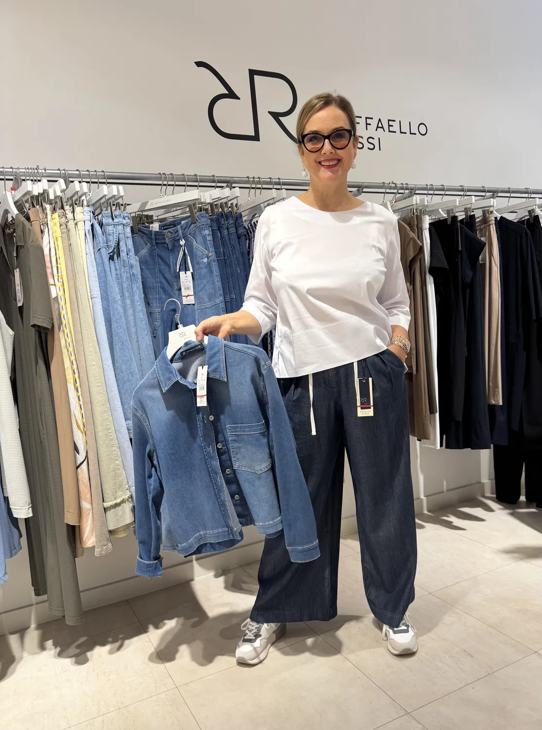 Abig-Moden - Modeboutique - Eine Frau in einem weißen Oberteil und einer dunkelblauen Hose posiert in einem Bekleidungsgeschäft und hält eine Jeansjacke vor einem Jeansständer hoch.