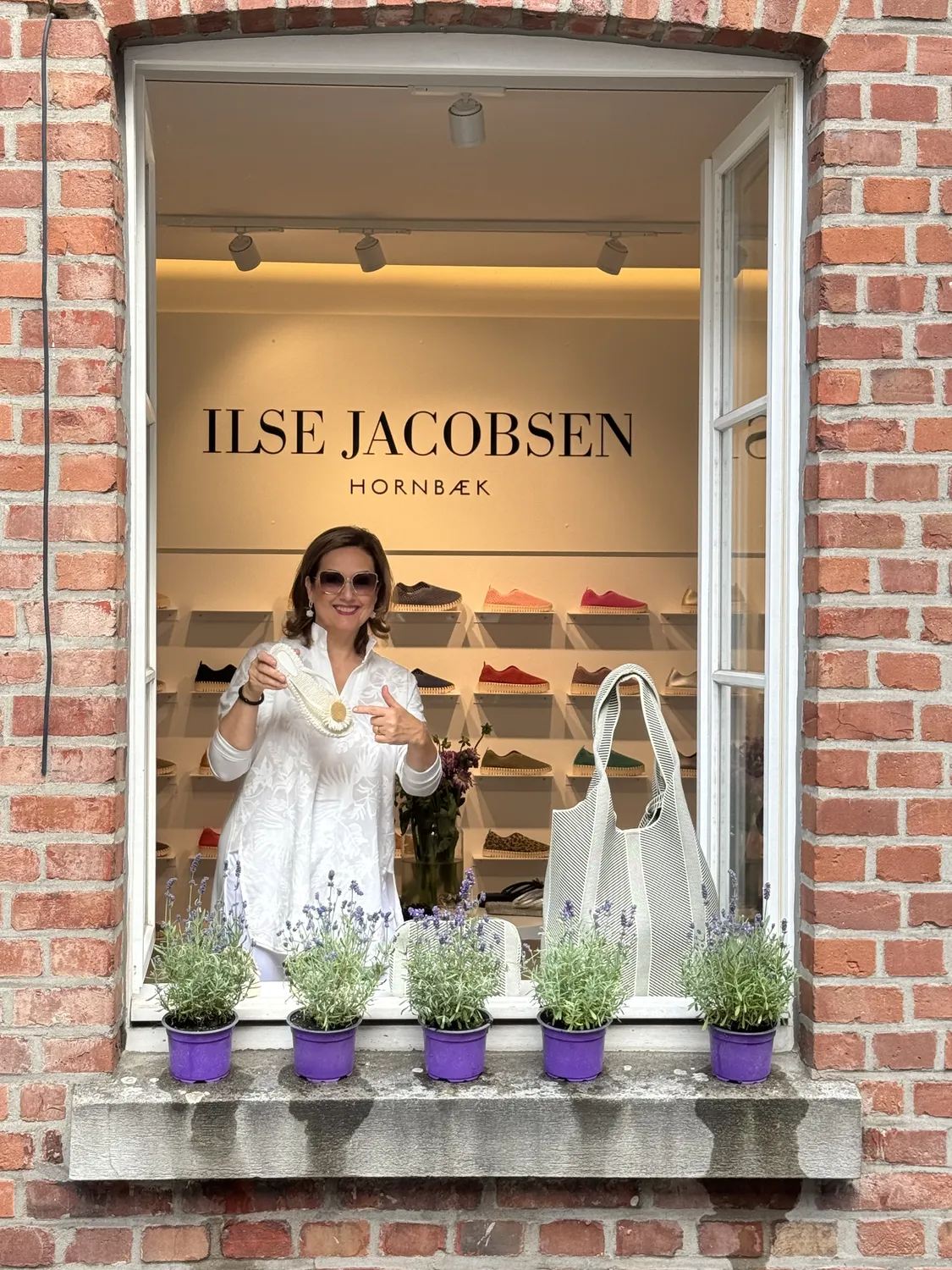 Abig-Moden - Modeboutique - Eine Frau lächelt aus dem Schaufenster einer Ilse Jacobsen Boutique, das von roten Backsteinen umrahmt und mit fünf kleinen lila Topfpflanzen auf dem Fenstersims dekoriert ist.
