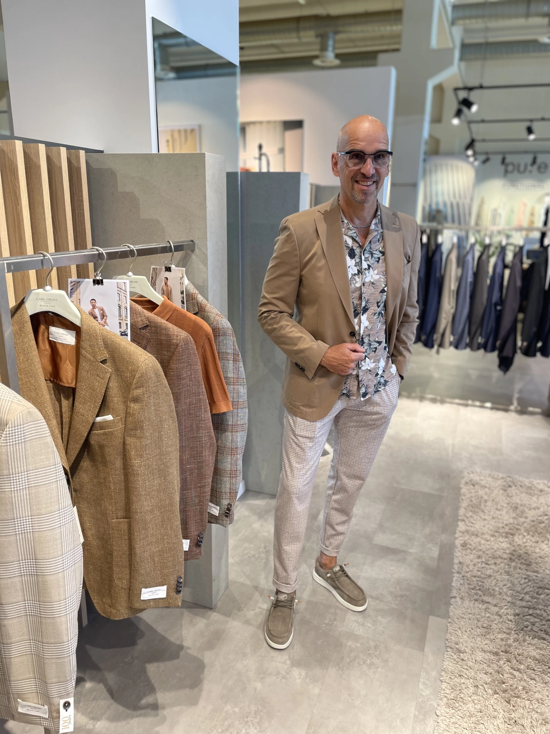 Abig-Moden - Modeboutique - Ein kahlköpfiger Mann, der einen beigen Blazer, ein Hemd mit Blumenmuster und eine helle gemusterte Hose trägt, steht in einer Bekleidungsboutique neben einem Ständer mit Sakkos.