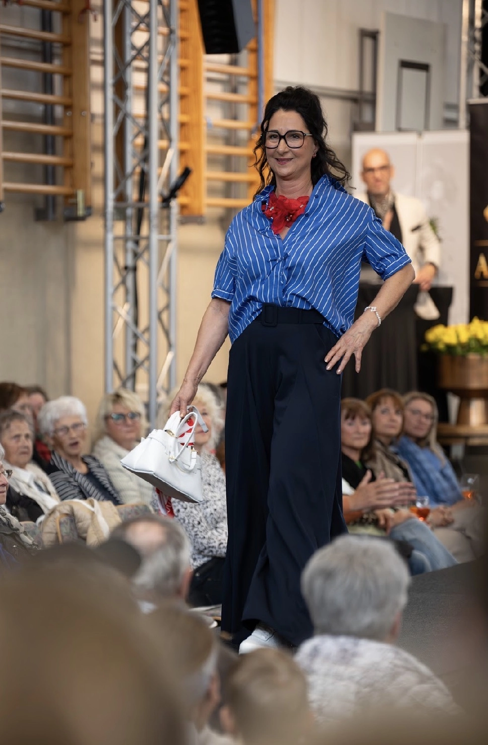 Abig-Moden - Modeboutique - Eine Frau in einem weißen Oberteil und einer dunkelblauen Hose posiert in einem Bekleidungsgeschäft und hält eine Jeansjacke vor einem Jeansständer hoch.