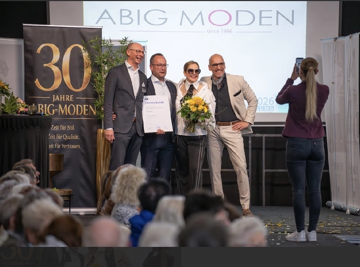 Abig-Moden - Modeboutique - Eine Frau in einem weißen Oberteil und einer dunkelblauen Hose posiert in einem Bekleidungsgeschäft und hält eine Jeansjacke vor einem Jeansständer hoch.